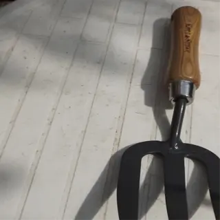 a garden fork with a wooden handle and metal tines, casting a shadow on a tiled surface