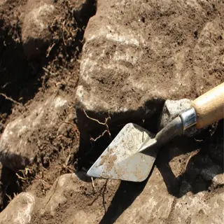 a hand trowel with a wooden handle rests on a rocky, uneven soil surface