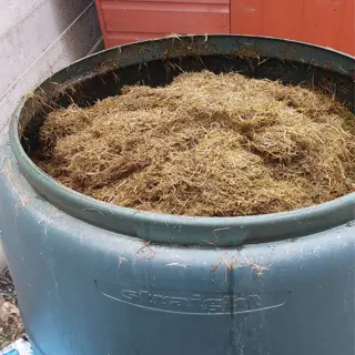 a large green compost bin filled with brown, dry grass clippings, placed on a concrete surface