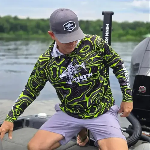a man wearing a stylish sun protection hat (gray cap) and neon fish-scale pattern shirt sits on a minn kota boat, showcasing shredfin fishing branding