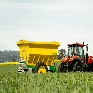 a red tractor pulling a yellow fertilizer spreader through a green crop field