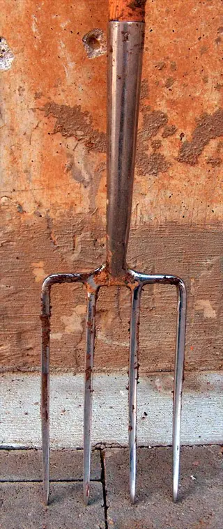 a weathered garden fork tool with wooden handle and metal tines rests against a rusty textured wall