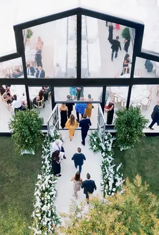 aerial view of a sweetheart patio wedding event with guests in formal attire entering a modern glass venue, floral-lined pathway, and elegantly set tables