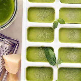 basil pesto ice cubes in white tray with fresh basil leaves, parmesan cheese, and grater - ready for freezing and later use