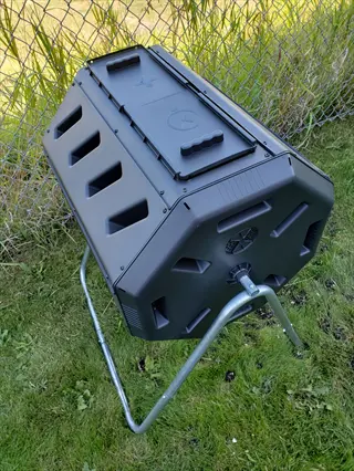 black barrel-style tumbling compost bin on a metal stand with ventilation slots, positioned in a grassy yard near a chain-link fence