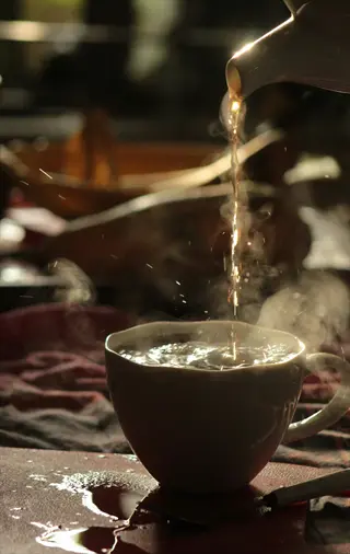 black tea cup pouring from teapot into ceramic cup with rising steam on cloth-covered table