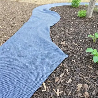 blue woven landscape fabric covering mulch path in a garden, with young plants and a tree trunk