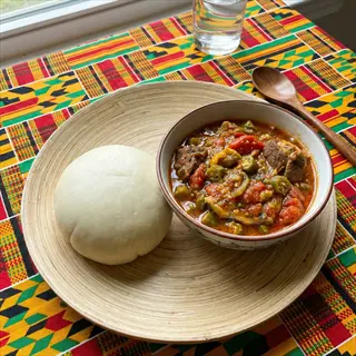 bowl of hearty okra stew with meat chunks and vegetables, served with a round dough on a woven plate over a colorful kente cloth-patterned table