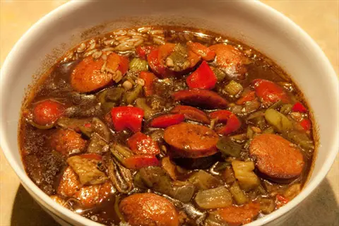 bowl of simmering okra gumbo with sliced sausages, bell peppers, and vegetables in a rich, dark roux-based stew