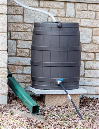 brown plastic rain barrel connected to green downspout and black hose, elevated on stone platform against stone house wall in residential garden