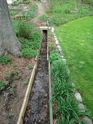 cedar raised bed drainage channel in garden landscape, directing water flow through wooden trench surrounded by lush plants, mulch, and lawn