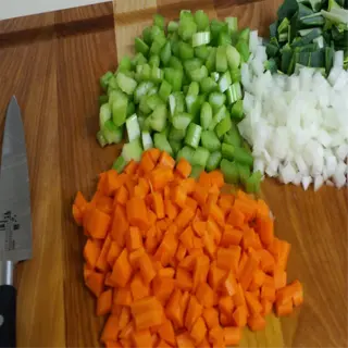 chopped vegetables including carrots, celery, onions, and greens on a wooden cutting board with a knife