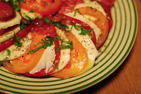 close-up of heirloom tomato caprese salad with red and orange tomatoes, fresh mozzarella, basil, and seasoning on a green-striped plate