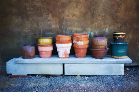 collection of terra cotta herb pots in varied sizes and glazes (gold, rust, teal) displayed on a concrete bench against a textured stone wall with gravel ground