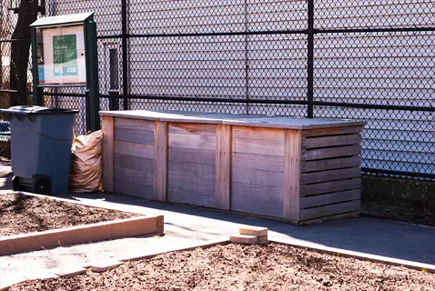 community garden compost: large wooden compost bin structure beside chain-link fence, with raised garden beds, trash bin, and bagged material in urban setting