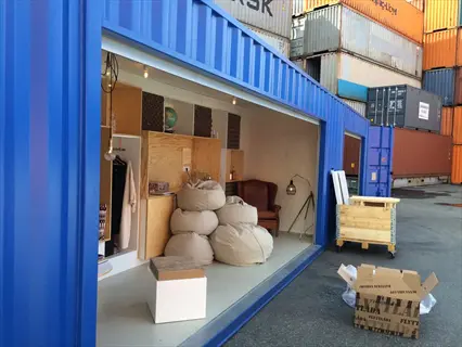 container adaptation: blue shipping container converted into a modular lounge space with bean bags, armchair, and interior lighting, surrounded by stacked cargo containers with visible text markings
