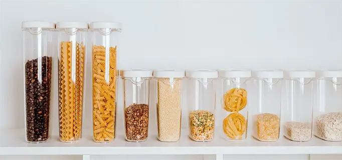 dried beans storage in clear containers organized on a white shelf, featuring legumes alongside other pantry staples like pasta and grains