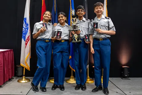 four individuals in blue cadet uniforms proudly holding awards on a stage with flags in the background