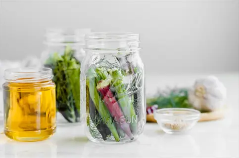 fresh okra refrigerator storage preparation: green and red okra pods in mason jars with dill, garlic, vinegar, and spices on marble surface