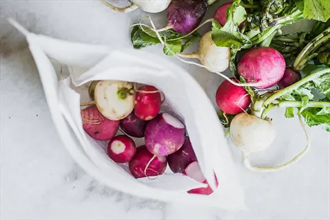 fresh radishes in a white cloth bag (demonstrating damp cloth radishes preservation method) with loose roots and greens on marble surface