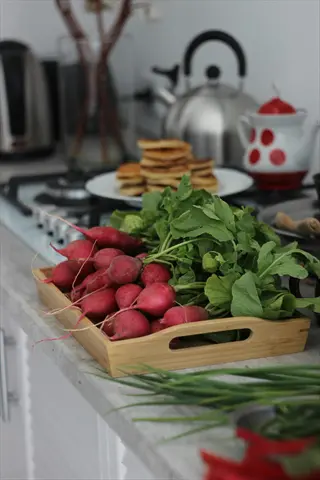 fresh radishes with green tops in a wooden tray on a kitchen counter; background features pancakes, a kettle, and kitchen appliances. a vibrant radishes kitchen counter harvest display