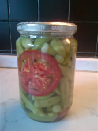 glass jar of pickled okra with tomato slice, visible text on lid ('gar...'), placed on a kitchen countertop with tiled backsplash