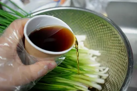 gloved hand pouring lemongrass syrup from a glass into a strainer of green onions during food preparation