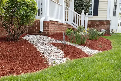 grass mulch garden: red mulch and white gravel landscaping with green grass, shrubs and daffodils near house porch with white railing