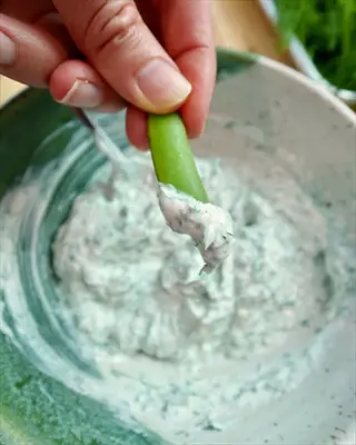 hand dipping cucumber into creamy tzatziki with dill, showcasing the yogurt-based dip with fresh herbs