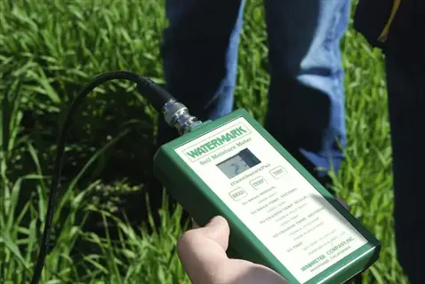 hand holding a watermark soil moisture meter in a grassy garden, measuring soil hydration with visible device text and people's legs in background