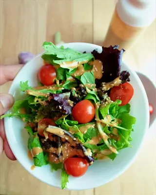 hand holding white bowl of mixed greens salad with cherry tomatoes and salad dressings. dressing bottle visible in background