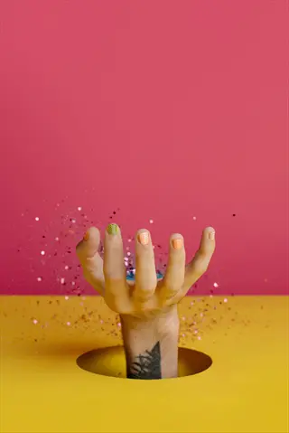 hand with multicolored fingernails (including pumpkin-textured orange nails) emerging from yellow circular cutout against pink background, surrounded by glitter confetti