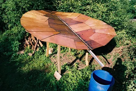 homemade rain collection system: large curved wooden roof structure supported by poles in a lush green setting, with blue barrel and firewood pile nearby
