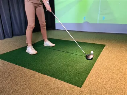 indoor golf setup with person preparing to swing at a golf ball (similar in size to a medium beet root) on artificial turf mat near simulator screen