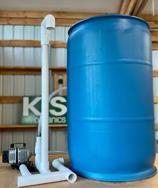 industrial compost tea brewing setup: blue barrel with white pvc aeration tubing and air pump. background shows 'k&s organics' branded sign in a barn setting