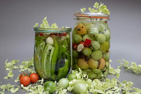 jar pickled green tomatoes with spices and herbs, alongside a jar of pickled green peppers and red chilies, with fresh tomatoes and white hydrangea flowers on gray surface