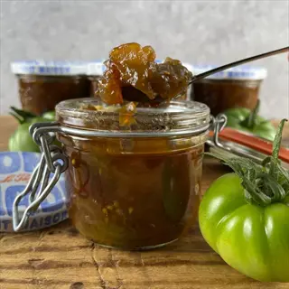 jars of green tomato chutney with spoon sample, fresh green tomatoes, and labeled jar lid ('maison') on wooden table