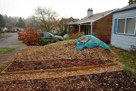 large pile of arborist wood chips covered with tarp in a residential front yard, with raised garden beds and houses on a misty day