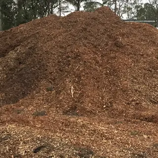 large pile of composted pine bark mulch in an outdoor setting. the dark brown, decomposed organic material forms a textured mound under overcast skies, with trees visible in the background