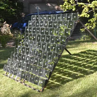 large sloped garden mirror array with grid of square mirrors in a grassy area, surrounded by trees and a blue vehicle in the background