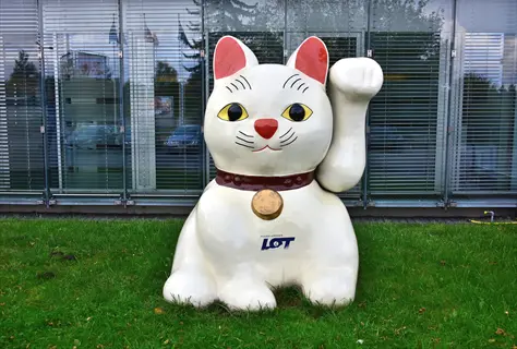 large white maneki-neko maskotka (lucky cat) statue with raised paw, red inner ears, and 'lot' text on its collar. displayed outdoors on grassy area in front of a modern glass building