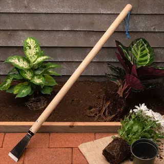 long-handled root harvesting tool with wooden handle and metal blade on brick surface, surrounded by potted plants against a wooden wall