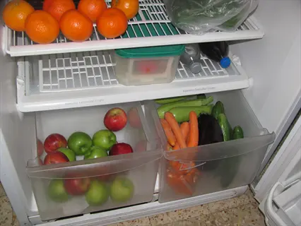 open refrigerator with carrots stored in the crisper drawer, alongside apples, zucchinis, and eggplants, with oranges on the shelf above