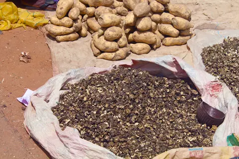 open sack of dried okra slices at a market stall, surrounded by piles of fresh potatoes and other dried produce on burlap sacks and cloth