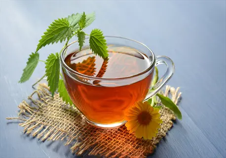 oregano herbal tea in a clear glass cup, garnished with fresh green leaves and a yellow flower, resting on a burlap mat against a soft blue background