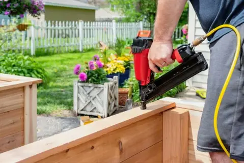 person assembling a pressure treated wood planter box using a pneumatic nail gun in a garden with flower pots and houses in the background