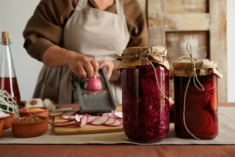 person slicing radishes for pickling preparation with two pickled radish jars (filled with red preserved vegetables) on a rustic kitchen table with ingredients