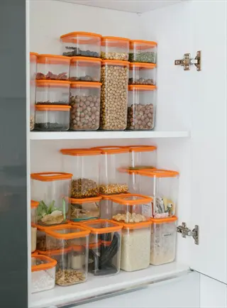 plastic seed storage cases in cabinet: multiple clear containers with orange lids organized on white shelves, storing various dried goods like seeds, grains, and nuts