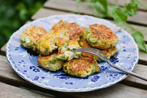 plate of golden okra fritters with visible okra pieces, arranged on a blue floral-patterned plate, one cut open with a fork, on a wooden table with greenery in background
