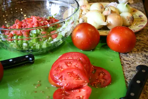 preparation of green tomato salsa: chopped green tomatoes and vegetables in a glass bowl, with whole tomatoes, onions, and knives on a green cutting board in a kitchen